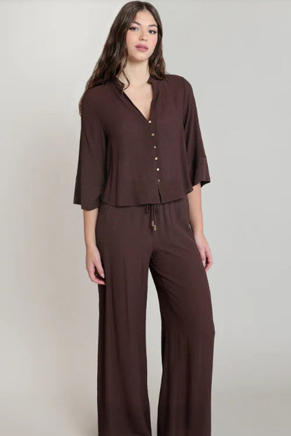 A woman modeling a dark brown two-piece set consisting of a short-sleeve button-front top and matching wide-leg pants, photographed against a plain light gray studio background.