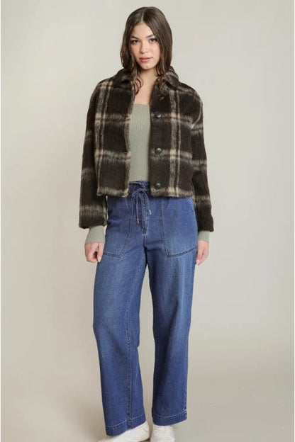 A model stands against a light beige studio background wearing a brown and black plaid cropped jacket over a sage green top, paired with blue wide-leg jeans and white sneakers.