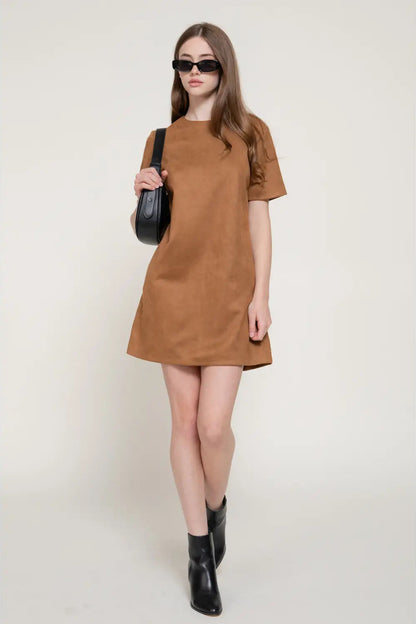 A woman modeling a short-sleeve brown dress with a relaxed, oversized fit, styled with black sunglasses, a black shoulder bag, and black ankle boots against a plain light beige background.