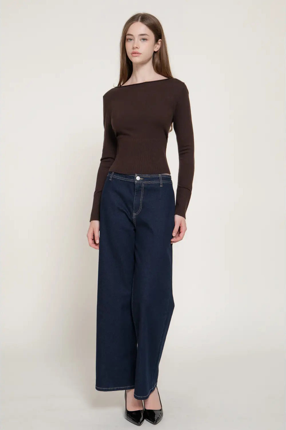 A model wearing a dark brown long-sleeve knit top and dark blue wide-leg jeans, standing against a plain light beige studio background.