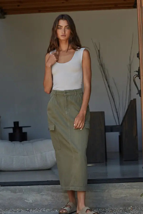A woman stands in a modern porch setting wearing an olive green high-waisted midi cargo skirt with side flap pockets, paired with a white tank top and flat sandals.