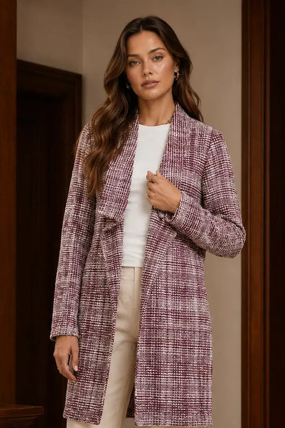 A woman wearing a textured, long-sleeve tweed coat in a dusty purple and pink pattern over a white top and beige trousers, standing in a warm-toned indoor setting with dark wood doors.