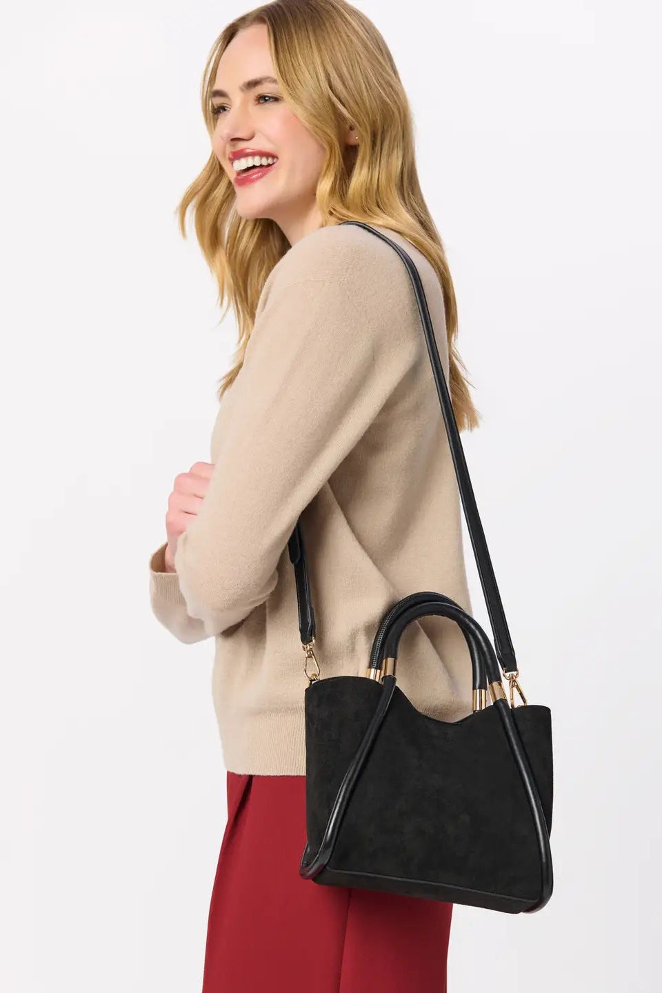 A woman wearing a beige sweater and red skirt carries a black suede-look shoulder bag with gold-tone hardware against a plain light gray studio background.