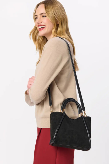 A woman wearing a beige sweater and red skirt carries a black suede-look shoulder bag with gold-tone hardware against a plain light gray studio background.