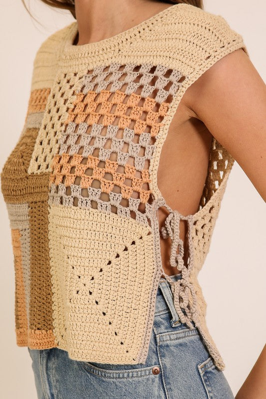 A close-up of a woman wearing a sleeveless crochet knit top with a textured patchwork design in beige, tan, and peach tones, paired with blue jeans.