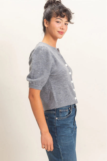 A woman wearing a gray short-sleeve knit cardigan with white buttons and blue jeans, standing against a plain light beige background.