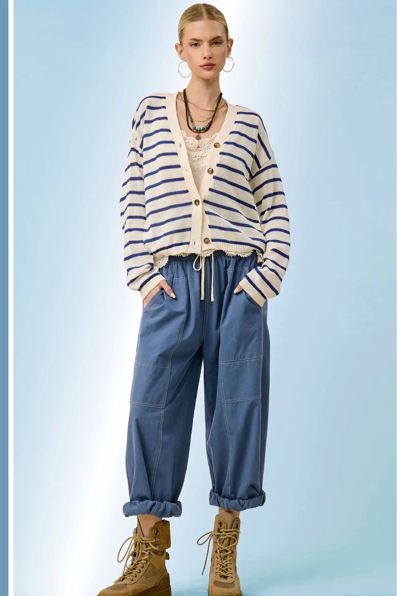 A model stands against a light blue studio background wearing a cream and navy blue horizontally striped cardigan over a cream top, paired with loose blue pants and tan lace-up boots.