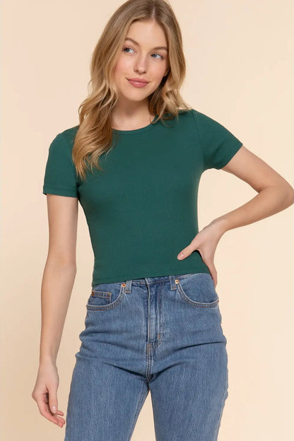 A woman modeling a fitted short-sleeve crewneck t-shirt in forest green, paired with high-waisted blue jeans, against a solid light beige background.