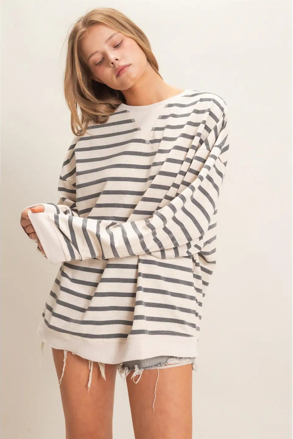 A woman wearing an oversized long-sleeve striped sweater with horizontal gray and off-white stripes, paired with frayed denim shorts, against a plain light beige studio background.