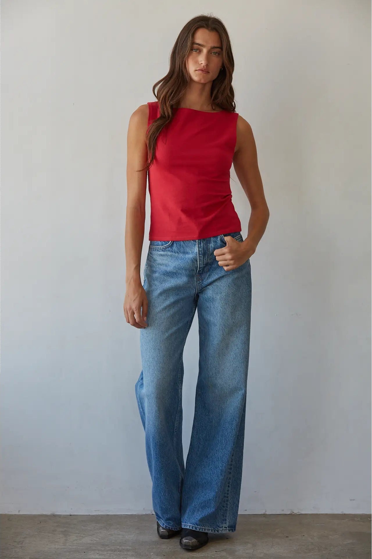 A woman modeling a sleeveless red top and wide-leg blue jeans against a plain light gray wall.