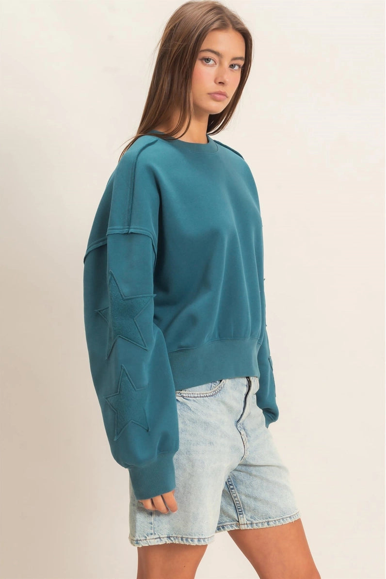 A woman modeling a teal crewneck sweatshirt with star-patterned sleeve embroidery, paired with light-wash denim shorts, against a plain beige background.