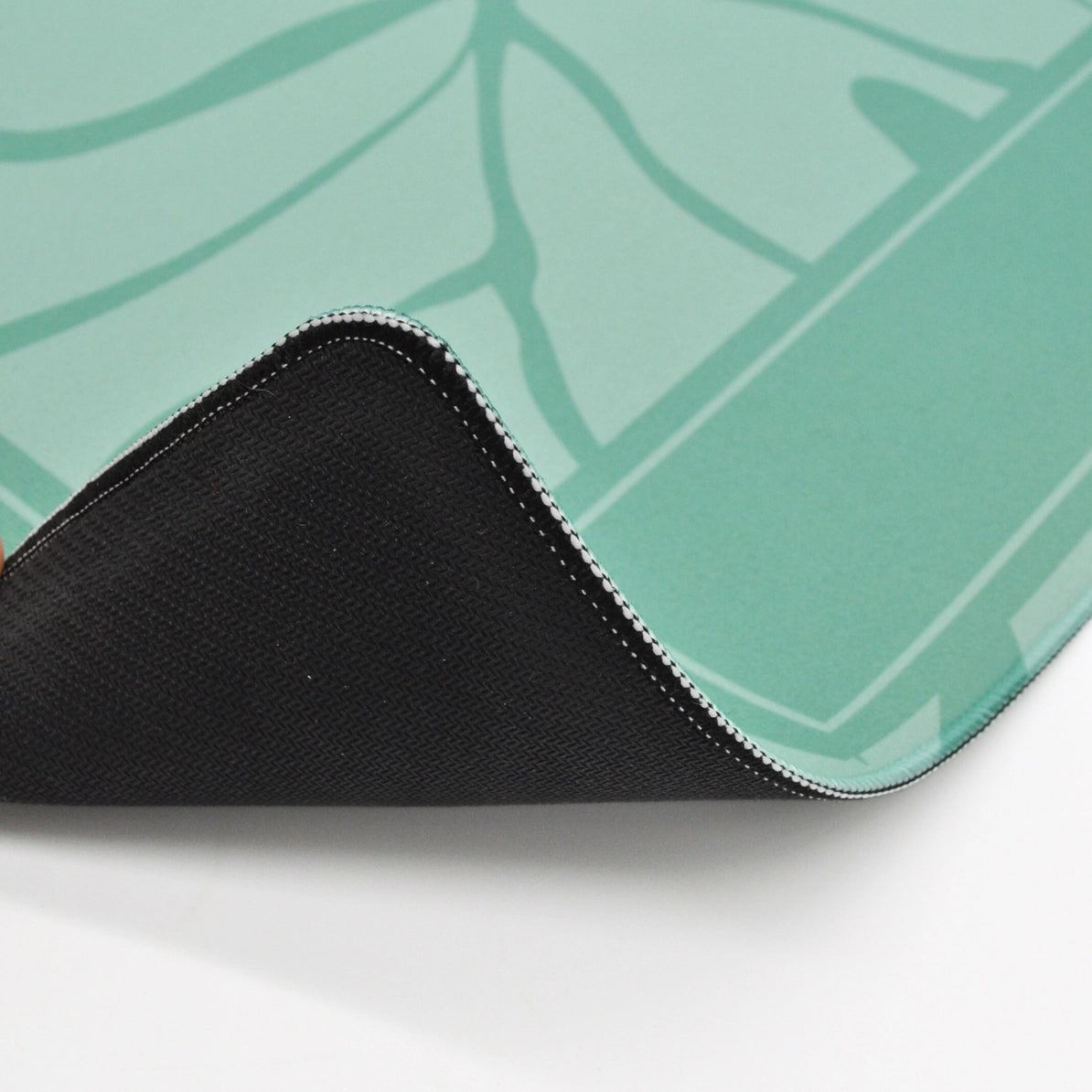 A close-up of a gaming mouse pad showing its black textured underside and a mint green top surface with a subtle leaf pattern, folded over on a white surface.