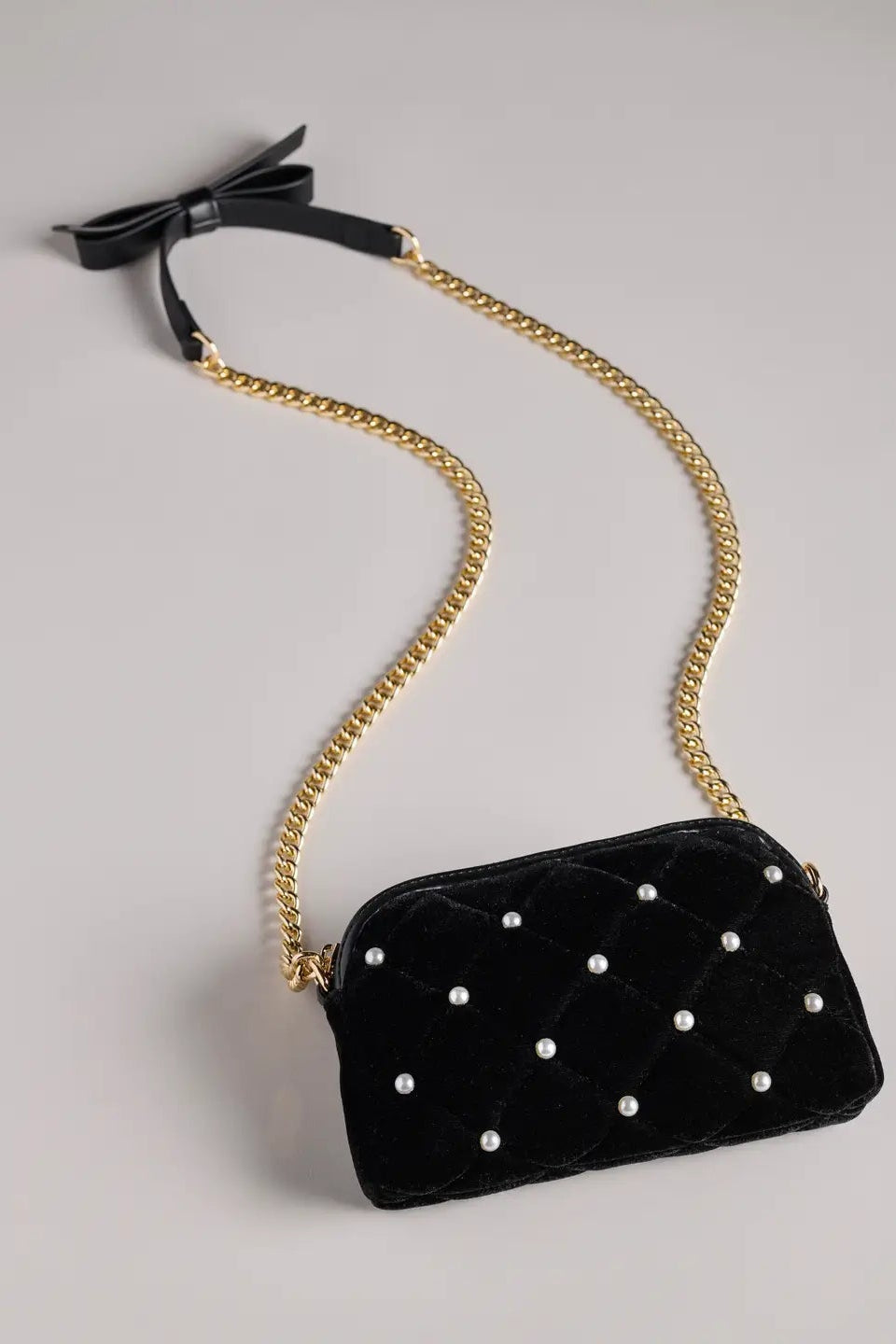 A black velvet crossbody bag with a quilted diamond pattern, adorned with small white pearls, featuring a gold chain strap and a black ribbon bow at the top, set against a plain light gray background.