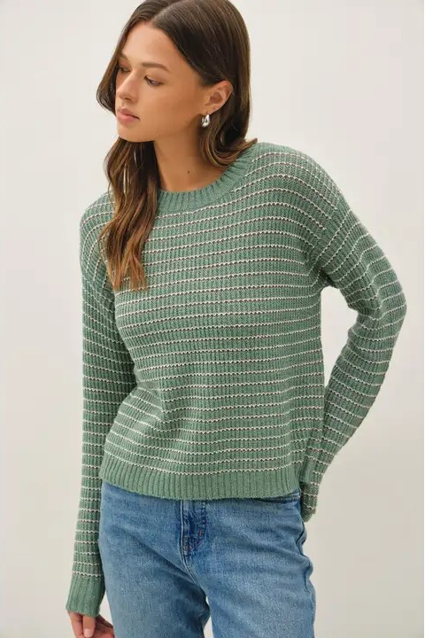 A woman wearing a sage green knit sweater with a textured white grid pattern, paired with blue jeans, photographed against a neutral light gray background.