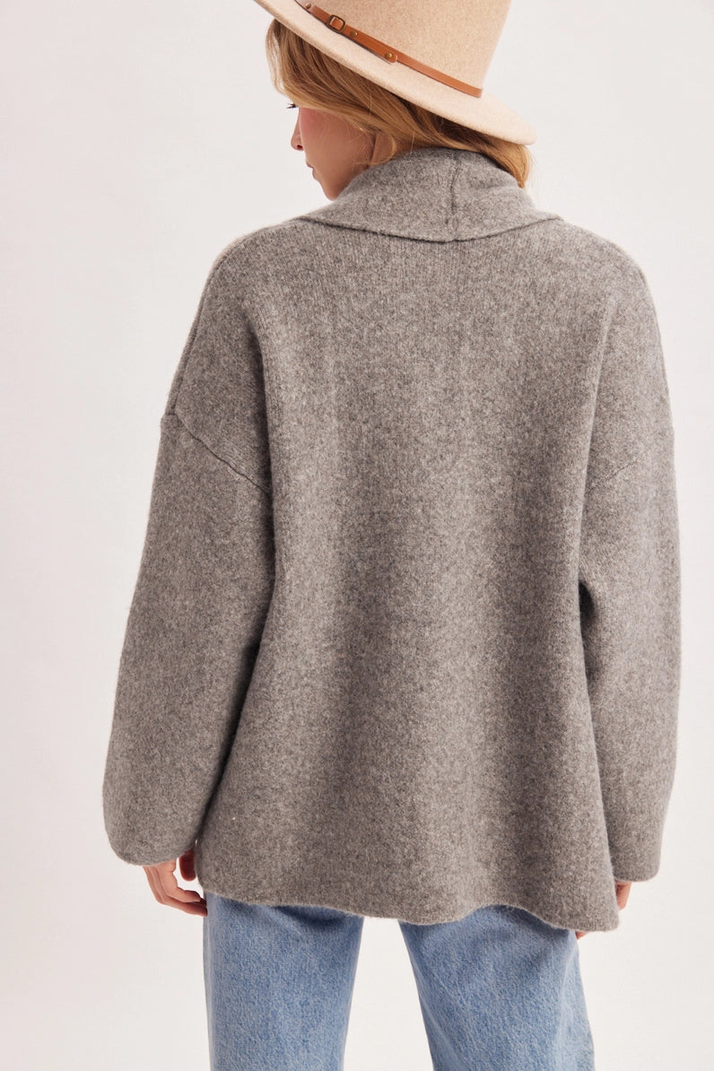 A person shown from the back wearing an oversized heather gray knit sweater with a wide collar, paired with light blue jeans and a beige hat, against a plain light background.