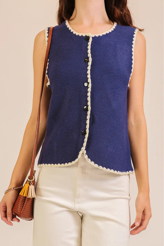 A woman wearing a sleeveless navy blue knit vest with cream scalloped trim and decorative buttons, paired with cream pants and carrying a brown shoulder bag.