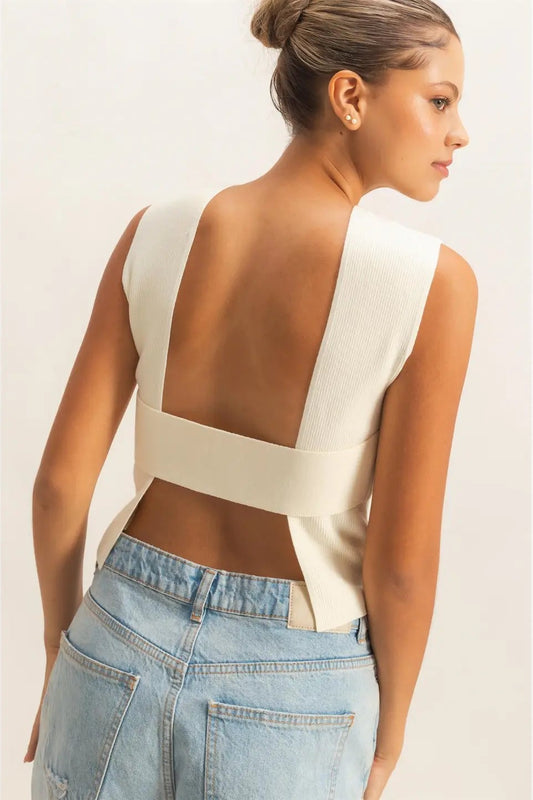 Woman wearing a white sleeveless top with cut-out design and blue jeans on a plain background