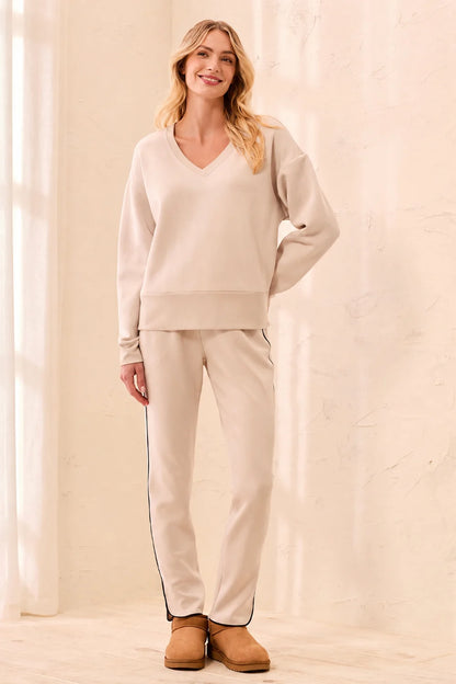 A woman modeling a matching beige loungewear set consisting of a V-neck long-sleeve sweatshirt and straight-leg sweatpants with thin dark side stripes, paired with tan boots, in a brightly lit cream-colored room.