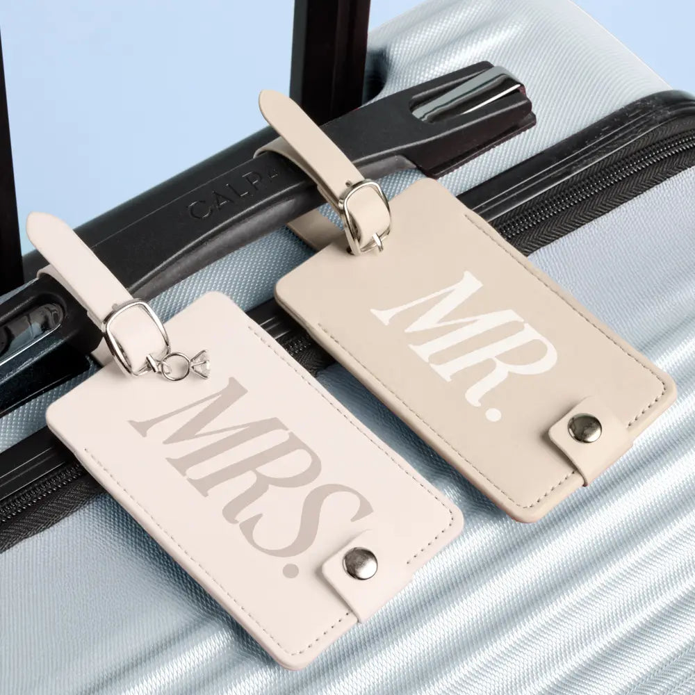 Two beige faux-leather luggage tags with silver metal clips, one printed with 'M.R.