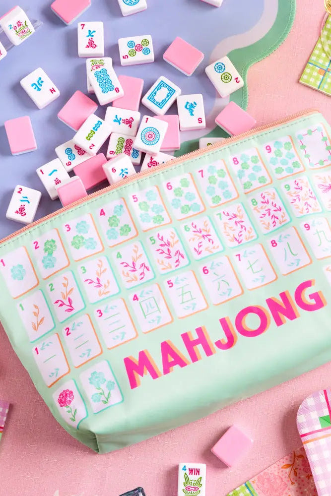 A mint green zippered pouch decorated with a Mahjong tile pattern and the word 'MAHJONG' in pink, surrounded by scattered pink and white tile pieces on a pastel background.