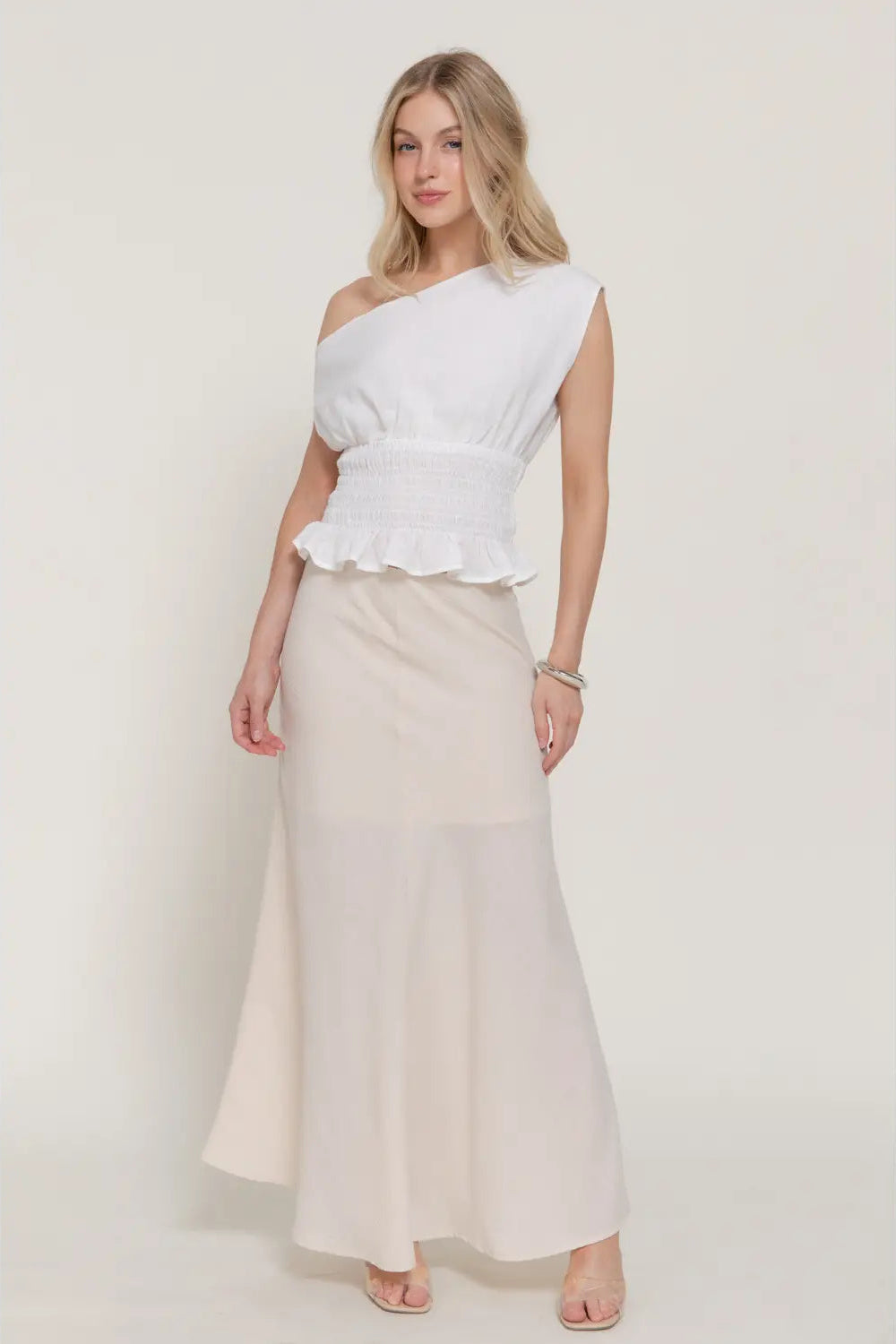 Woman wearing a white one-shoulder top and cream skirt on a plain background