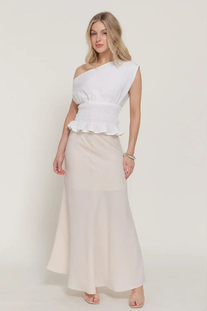 Woman wearing a white one-shoulder top and cream skirt on a plain background