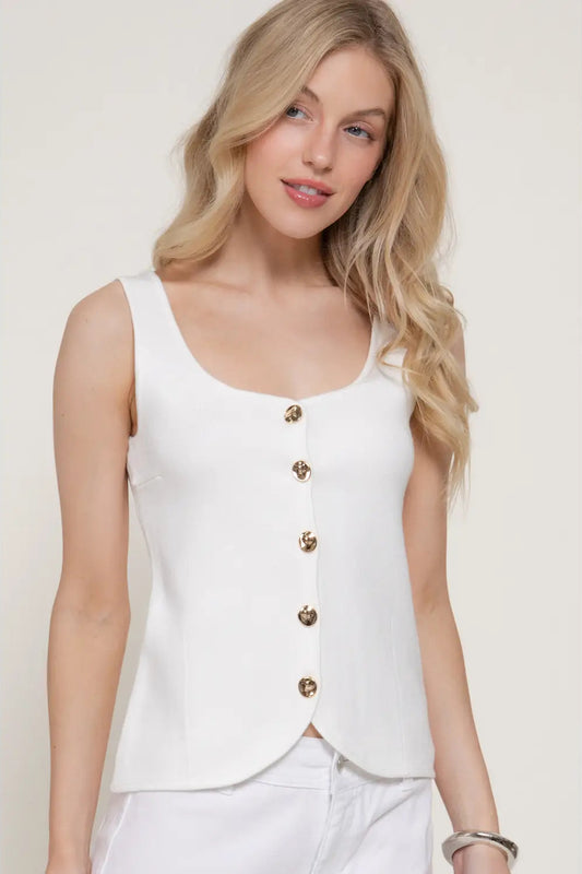 Woman wearing a white sleeveless top with gold buttons on a plain background