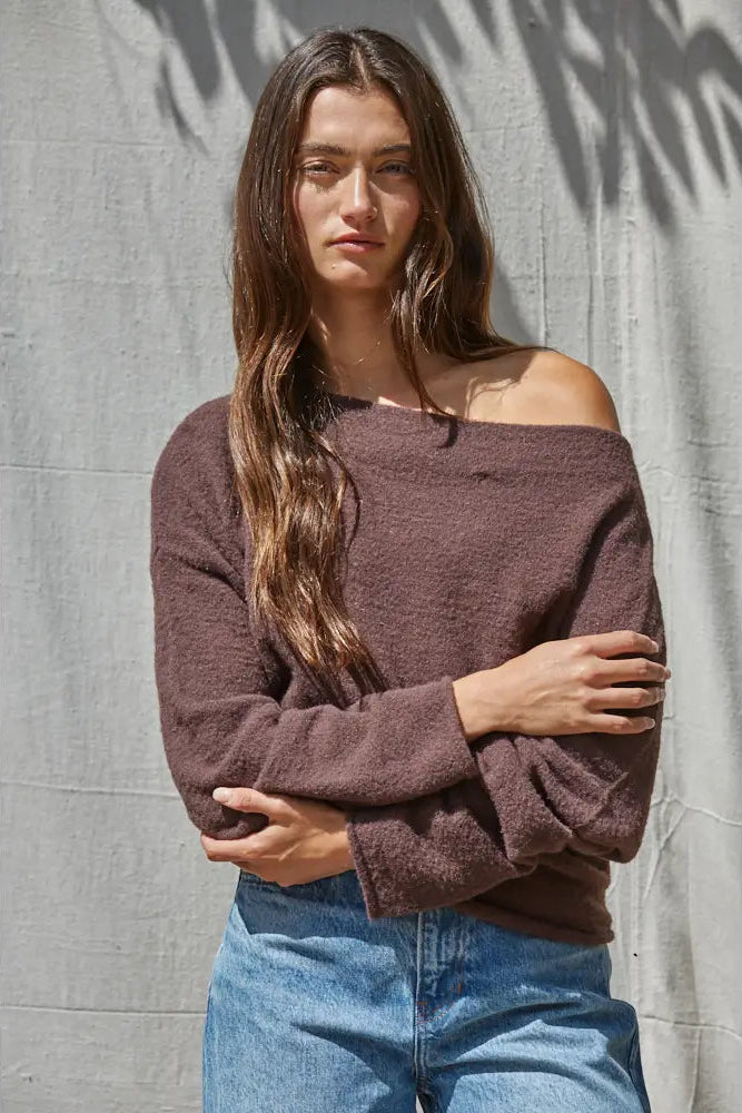 A woman wearing a fuzzy off-the-shoulder long-sleeve sweater in a dusty mauve-brown color, paired with blue jeans, standing against a textured gray wall with leaf shadows.