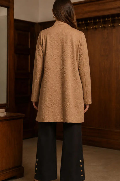A woman viewed from behind wearing a camel-colored textured jacket and black wide-leg trousers with gold buttons, standing in a room with dark wood paneling.