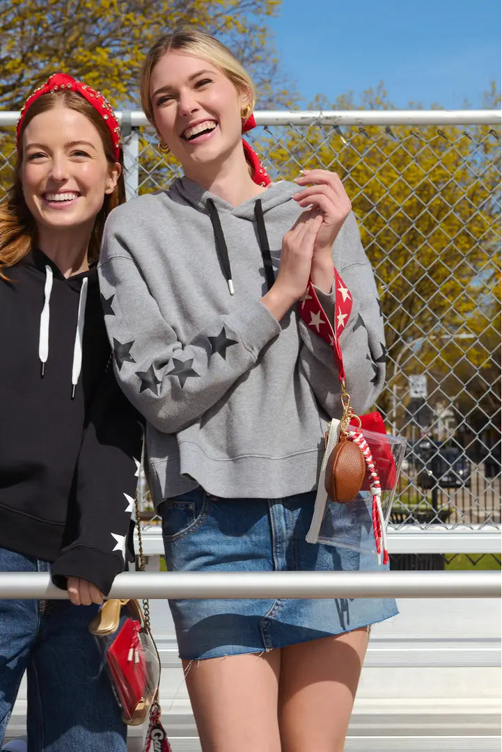 Two women smiling and posing outdoors in front of a chain-link fence. The woman on the left wears a black hoodie with white stars on the sleeves and a red bandana, while the woman on the right wears a gray hoodie with black stars on the sleeves and a red-and-white patterned crossbody bag.