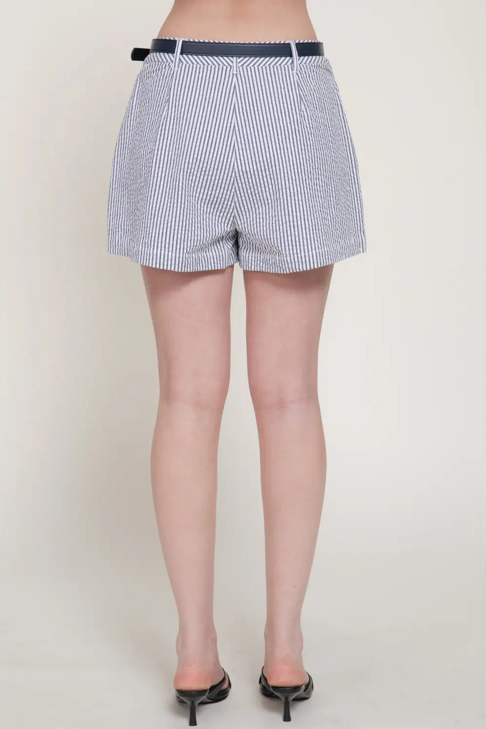A person shown from the back wearing high-waisted, blue and white vertically striped shorts with a thin dark belt, paired with black pointed-toe mule heels against a plain light beige studio background.