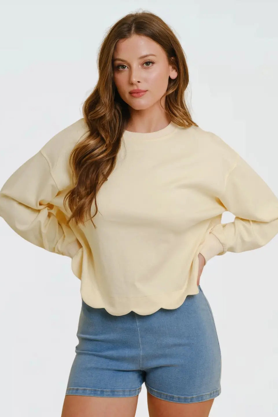 A woman modeling a pale yellow long-sleeve crewneck sweatshirt with a scalloped hem and balloon sleeves, paired with blue denim shorts, against a plain white studio background.