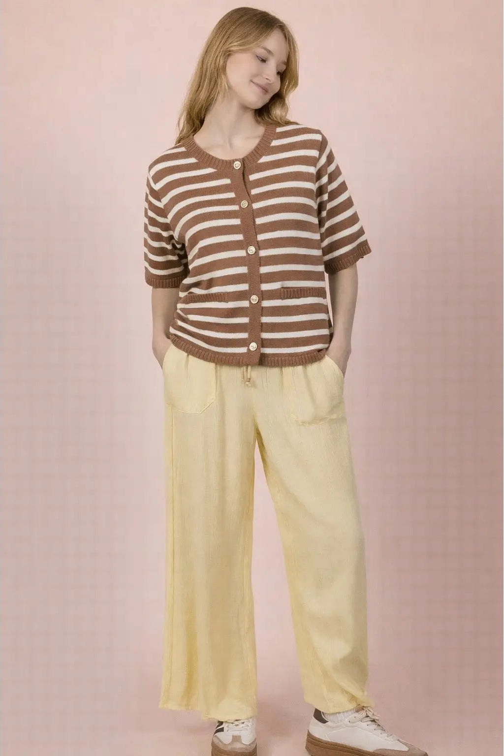 A woman modeling a brown and beige striped short-sleeve cardigan with buttons, paired with pale yellow wide-leg pants and white sneakers, set against a soft pink background.