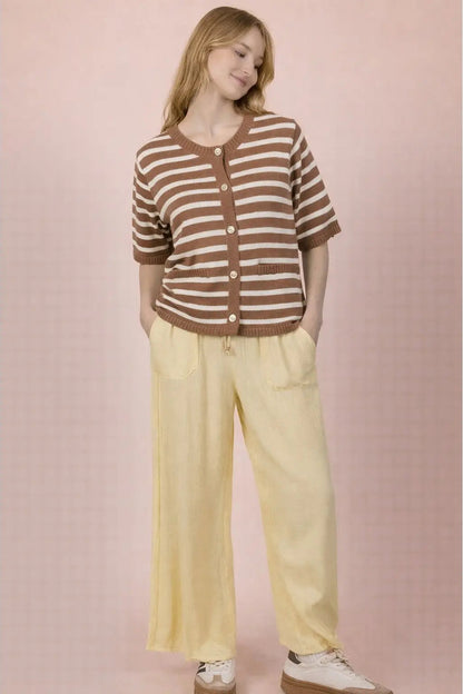 A woman modeling a brown and beige striped short-sleeve cardigan with buttons, paired with pale yellow wide-leg pants and white sneakers, set against a soft pink background.