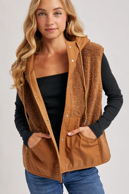 A woman wearing a brown sherpa-lined snap-button vest over a black long-sleeve top and blue jeans, photographed against a light gray studio background.