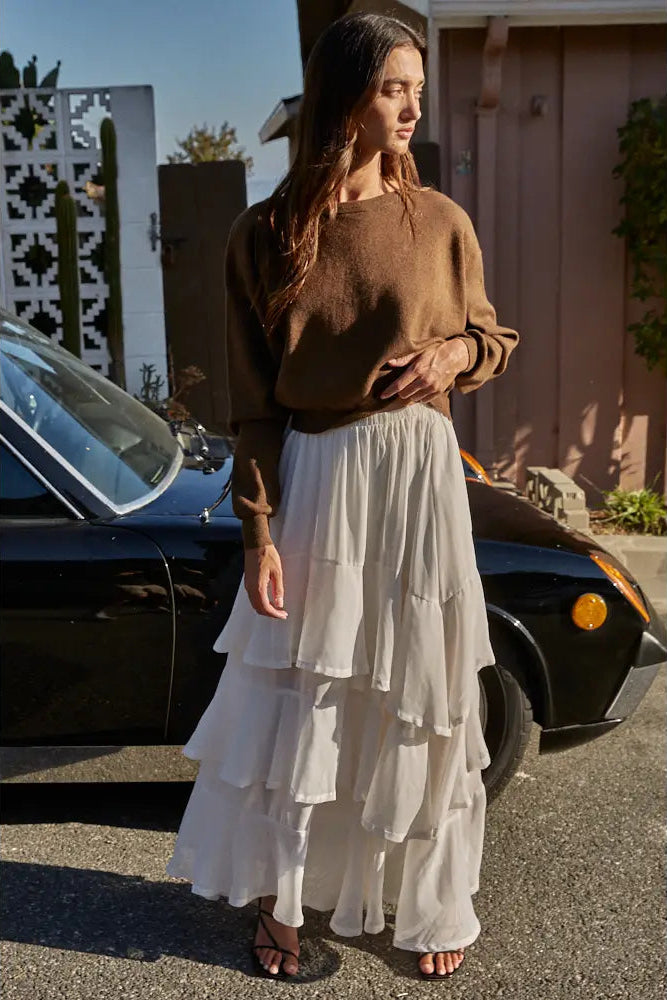 A woman stands outdoors in front of a black car and a house, wearing a brown long-sleeve knit sweater and a long white tiered skirt.