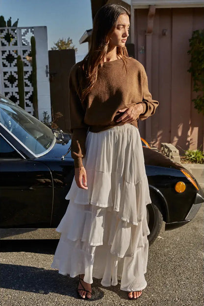 A woman stands outdoors in front of a black car and a house, wearing a brown long-sleeve knit sweater and a long white tiered skirt.