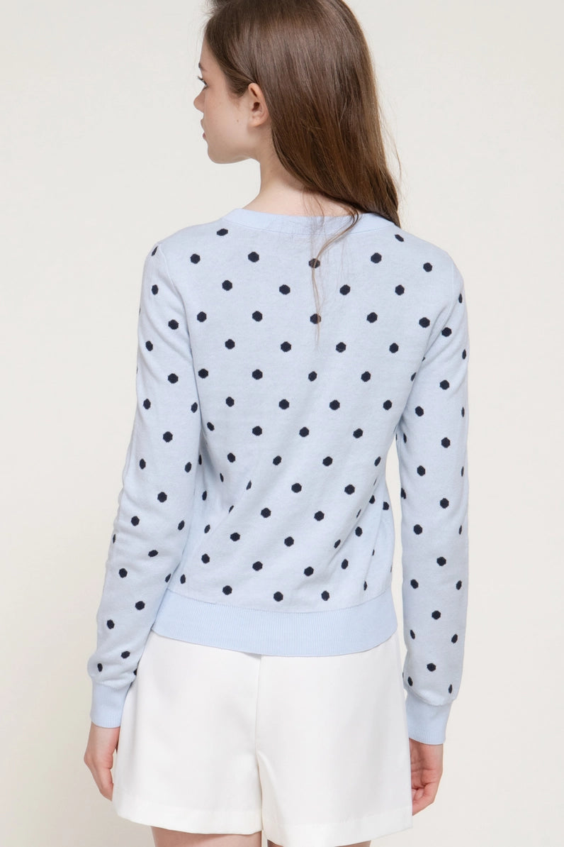 A woman shown from the back wearing a light blue long-sleeve top with black polka dots and white shorts against a plain light beige studio background.