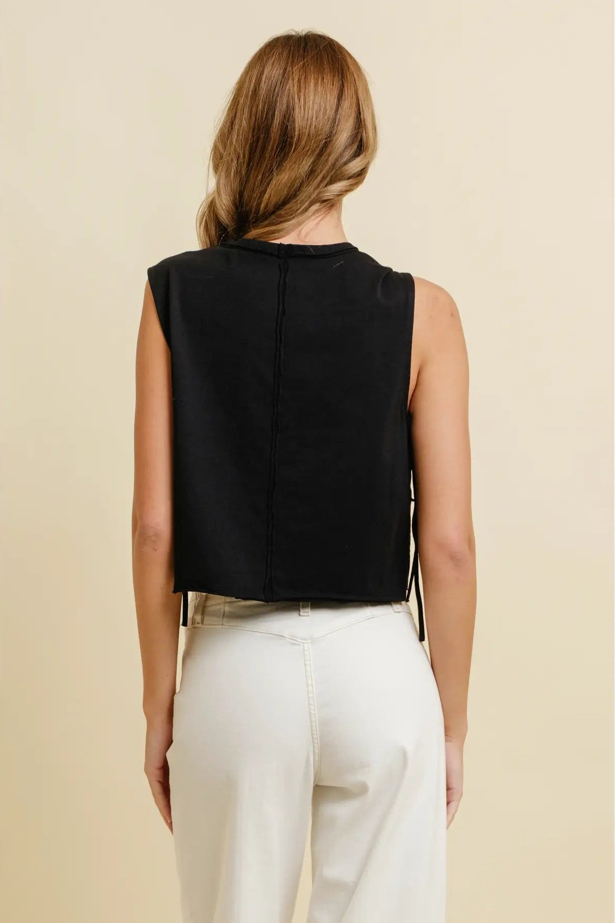 A woman shown from the back wearing a sleeveless black cropped top and cream-colored pants, set against a solid beige background.