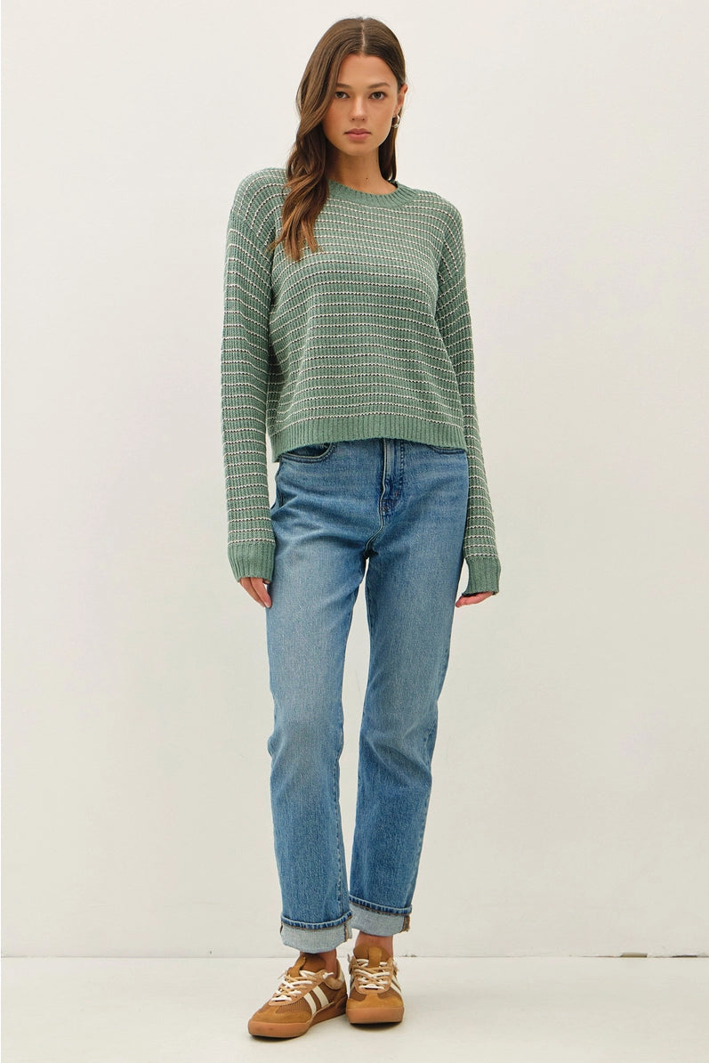 A woman modeling a green ribbed knit sweater with thin white stripes, paired with light blue jeans and brown sneakers, against a plain off-white studio background.