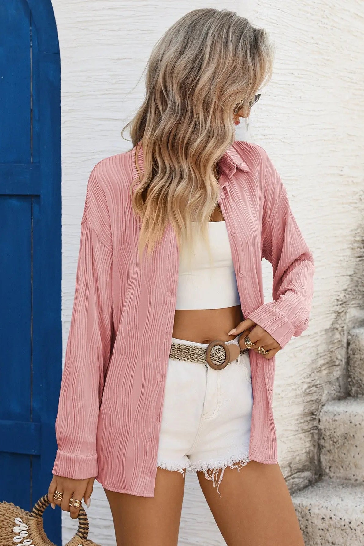 Woman wearing a pink shirt, white crop top, and shorts standing near a blue door.