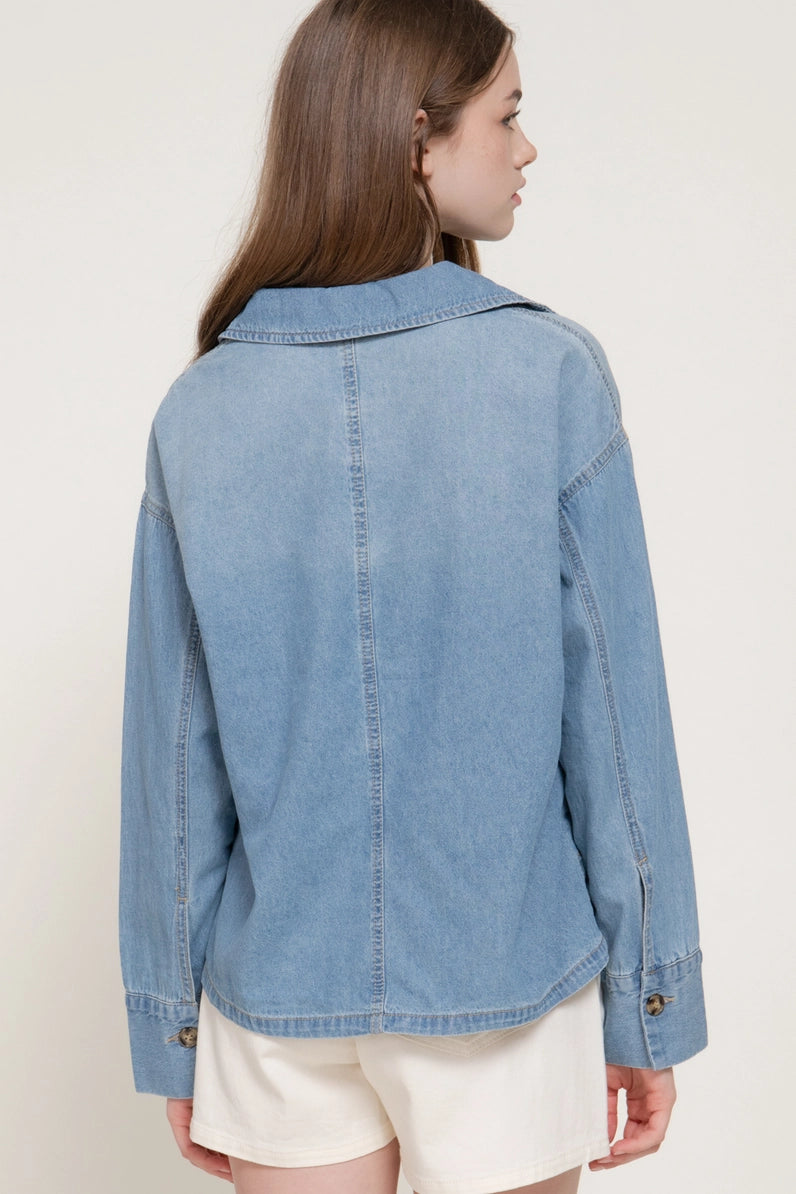 A woman shown from the back wearing a light blue denim jacket and cream shorts against a plain off-white studio background.