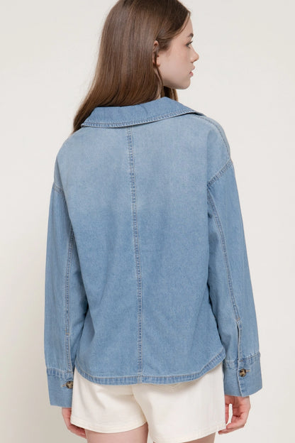 A woman shown from the back wearing a light blue denim jacket and cream shorts against a plain off-white studio background.