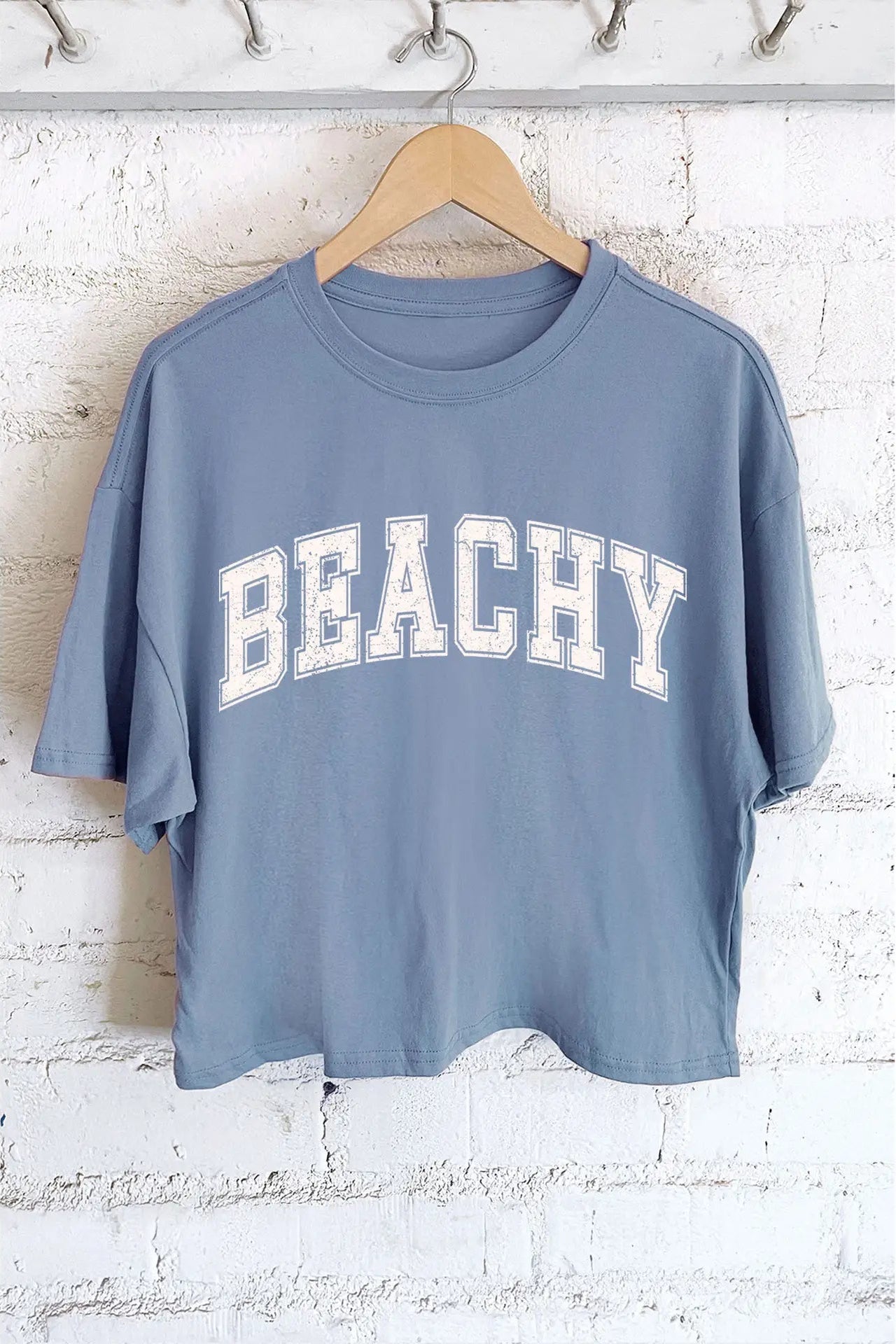 Blue t-shirt with 'BEACHY' text on a hanger against a white brick wall.