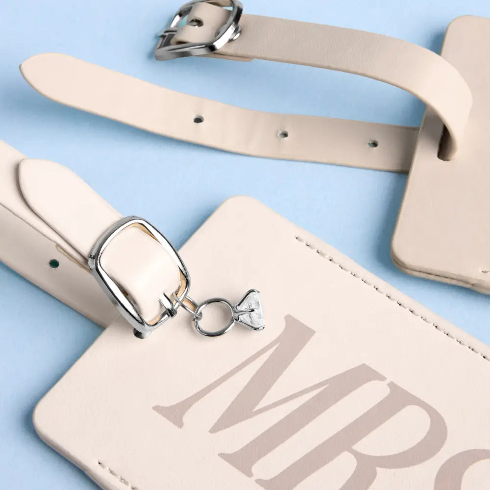 A close-up of a light beige leather luggage tag with a matching strap and silver-tone buckle, laid on a pale blue background. The tag has stitched edges and a small heart-shaped charm attached near the buckle.