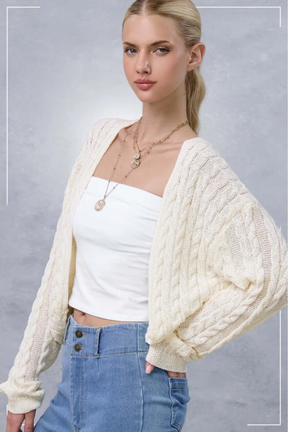 A blonde woman wearing a cream cable-knit cardigan over a white tube top and blue jeans, posed against a textured gray studio background.