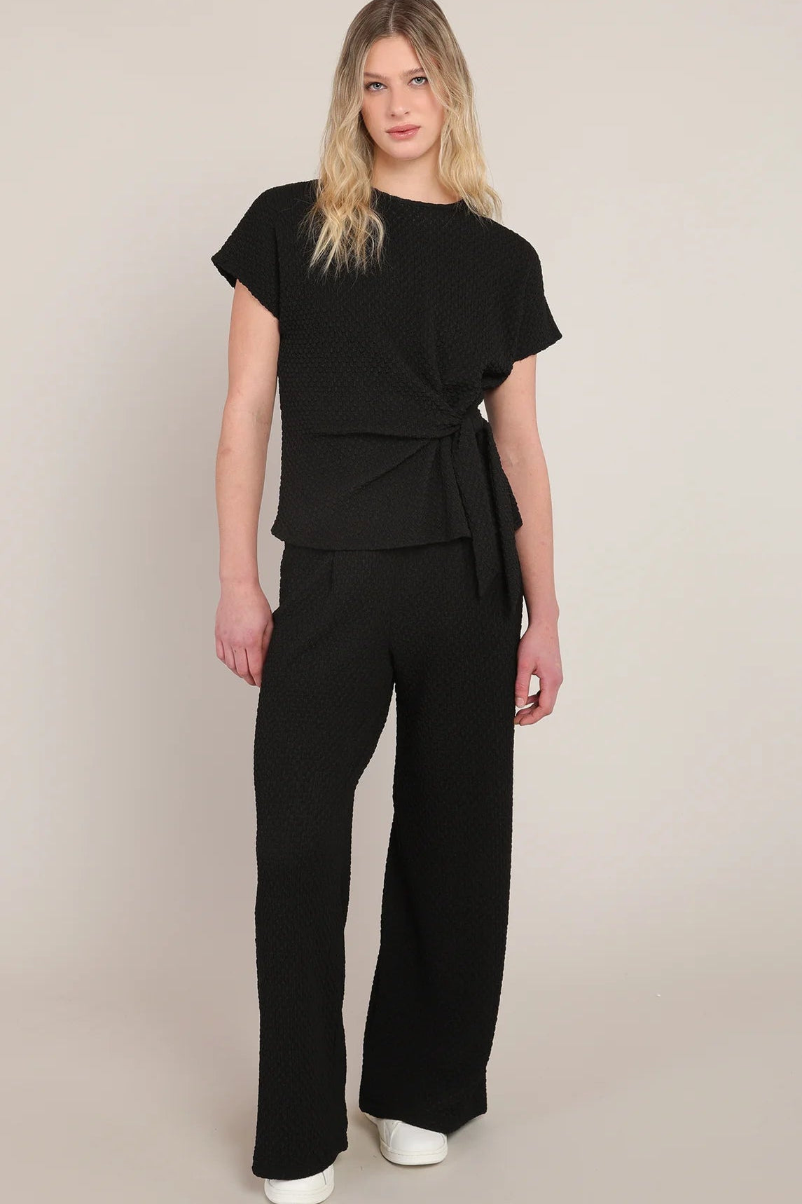 A woman modeling a black short-sleeve wrap-style top and matching wide-leg trousers against a plain beige studio background.