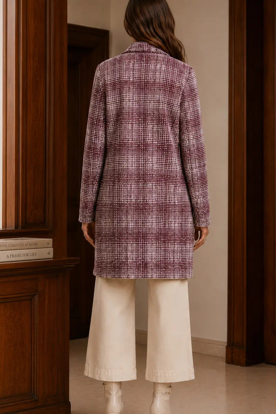 A woman viewed from behind wearing a long-sleeve plaid coat in dusty purple and white, paired with cream wide-leg trousers and white boots, standing in a room with dark wood paneling.