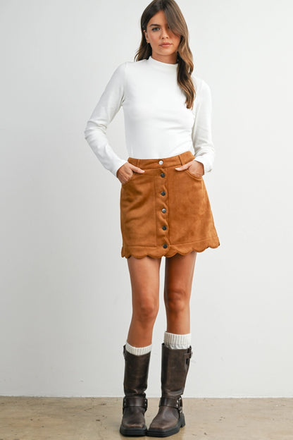 A woman modeling a brown suede button-front mini skirt with a scalloped hem, styled with a white long-sleeve mock-neck top and dark brown knee-high boots against a plain white studio background.