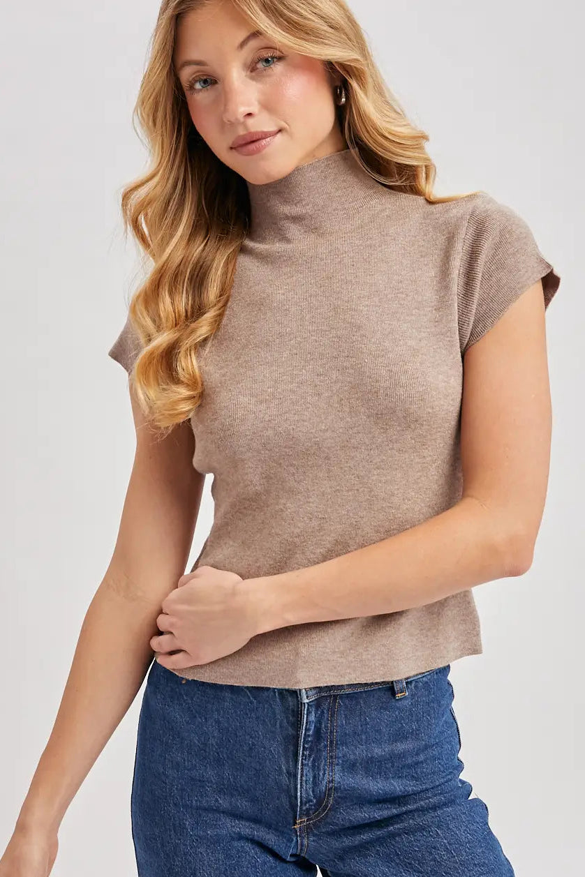 A woman wearing a taupe-brown short-sleeve mock neck knit top and blue jeans, photographed against a light gray studio background.